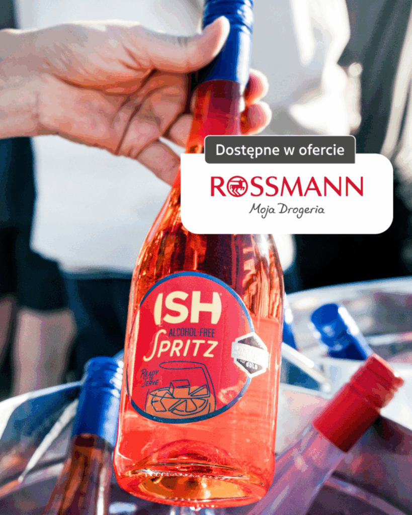 Butelka bezalkoholowego ISH Spritz trzymana w dłoni, dostępna w ofercie Rossmann, w letniej plenerowej scenerii z lodem i napojami.
